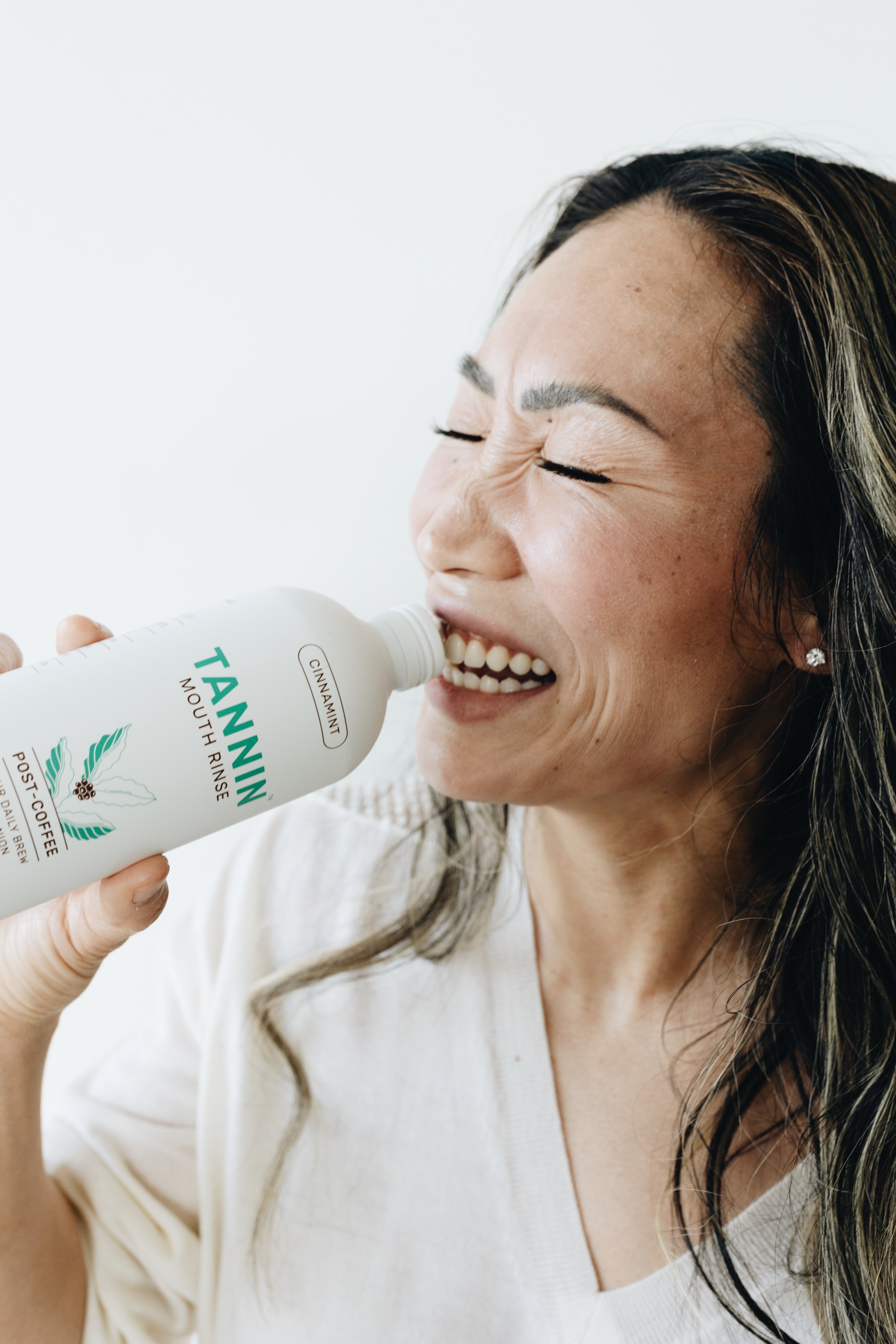 Smiling woman drinking from a bottle of Tannin Mouth Rinse in Cinnamint flavor, labeled for post-coffee use. She is holding the bottle close to her mouth and appears happy and refreshed. The product bottle displays Tannin’s branding and health-focused features.